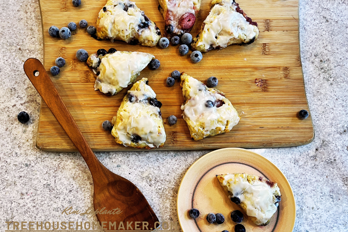 5 of the best mixed berry scones on a wood cutting board. one scone on a plate and a spatula to the size with blueberries scattered about